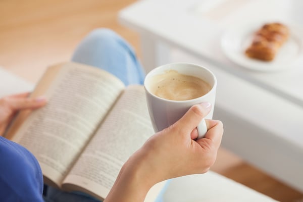 Woman sitting on the sofa reading a book holding her coffee mug in living room at home