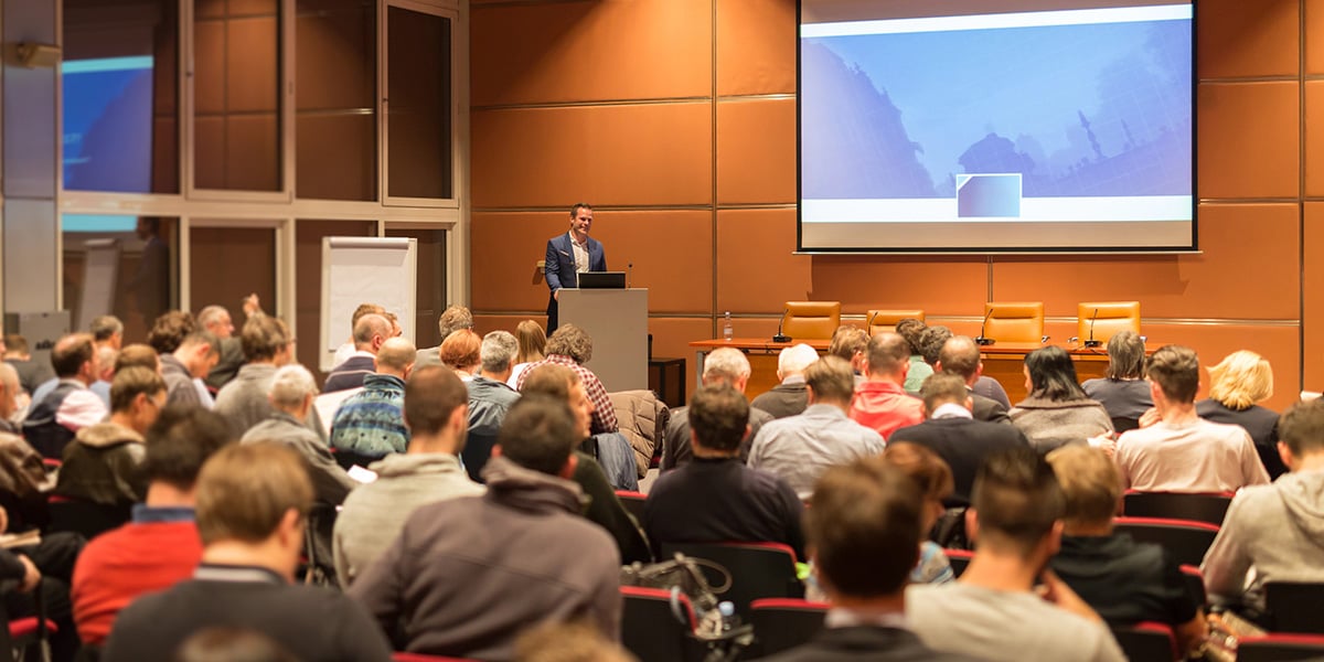 a speaker stands at a podium in front of a full event crowd in an auditorium